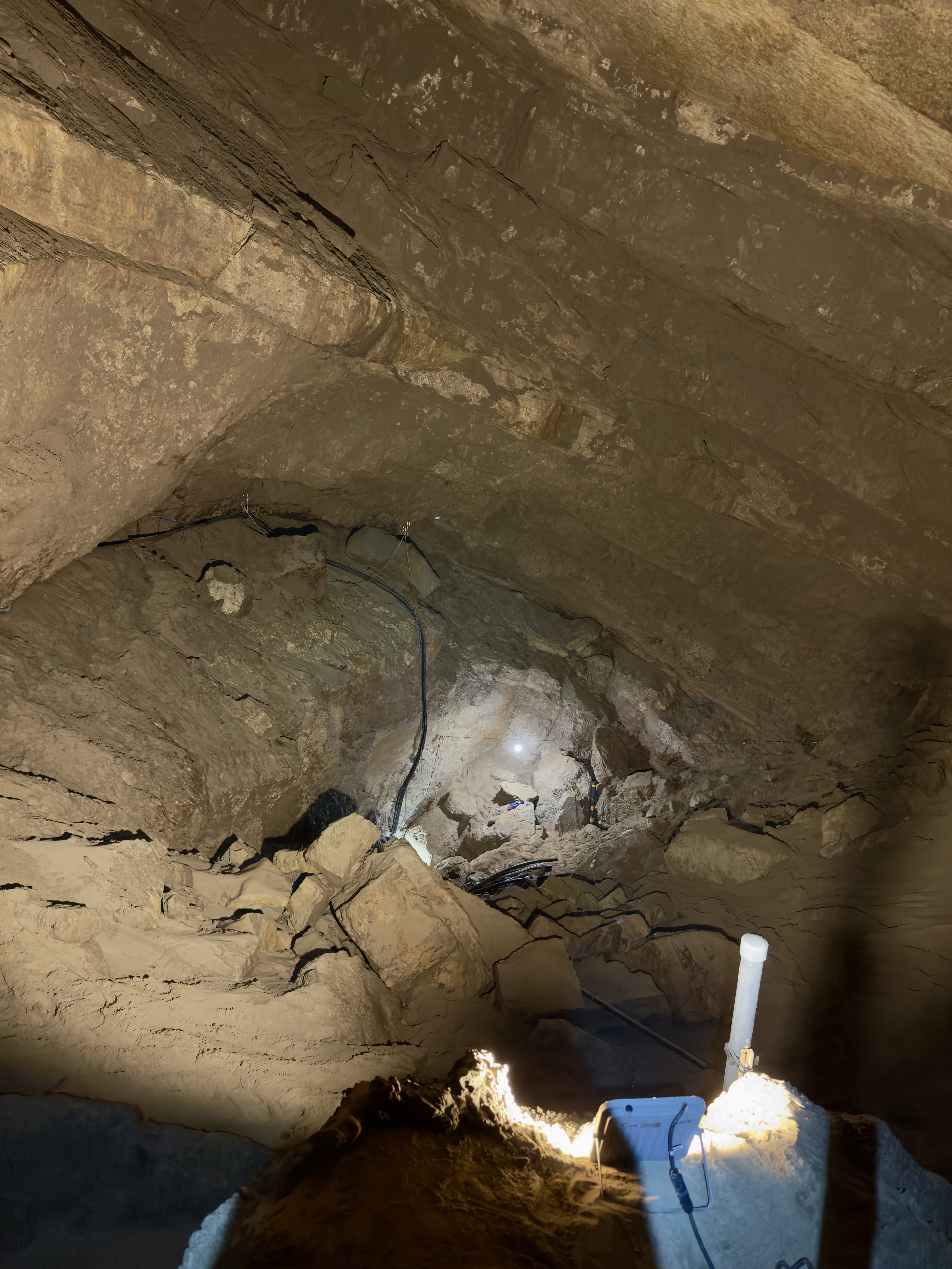 Vue d’une cavité rocheuse éclairée par des lampes, avec des blocs au sol et des câbles visibles le long des parois.