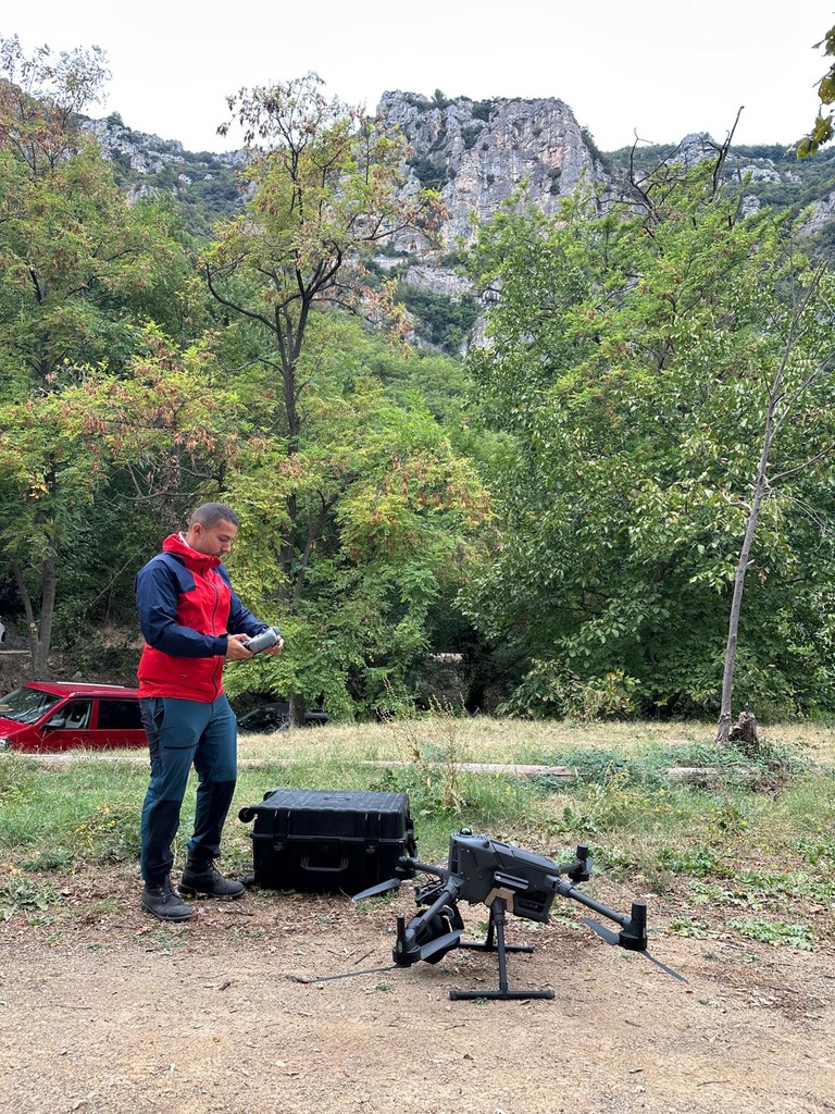 Un opérateur tient une radiocommande près d’un drone posé au sol ; une falaise et des arbres occupent l’arrière-plan.