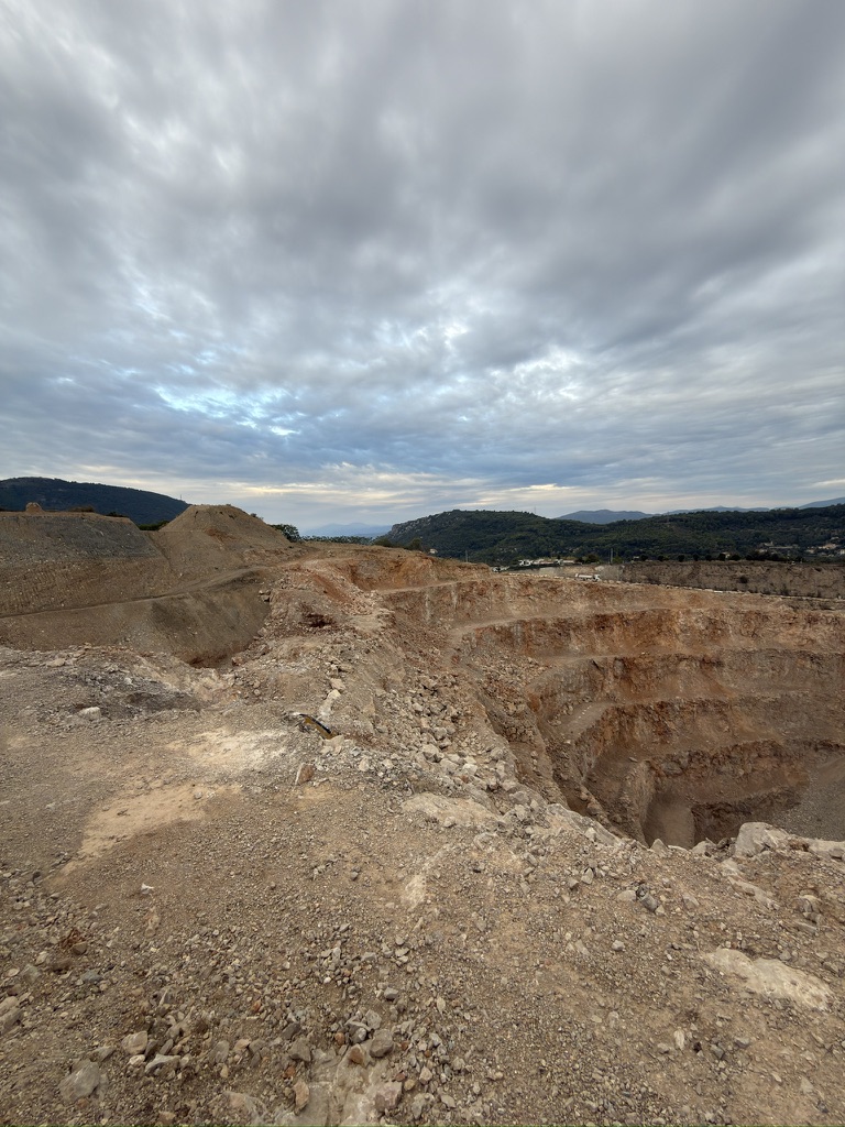 Vue de la carrière avec ses gradins rocheux, un sol caillouteux au premier plan et des collines à l’horizon.
