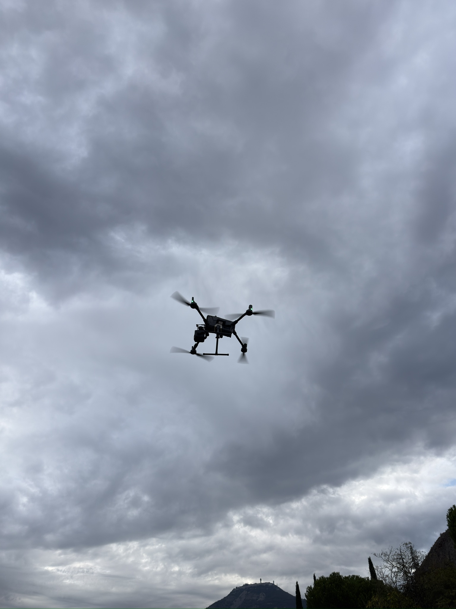 Drone photographié depuis le sol sous un ciel nuageux, avec une crête et de la végétation au bas de l’image.